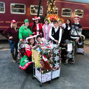 Group in festive attire poses by a decorated cart and old train.