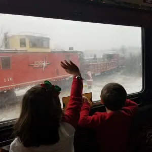 Two children waving at a red train through a window on a snowy day.