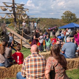 People sitting on hay bales facing a pirate ship with sails at an outdoor event under a cloudy sky.