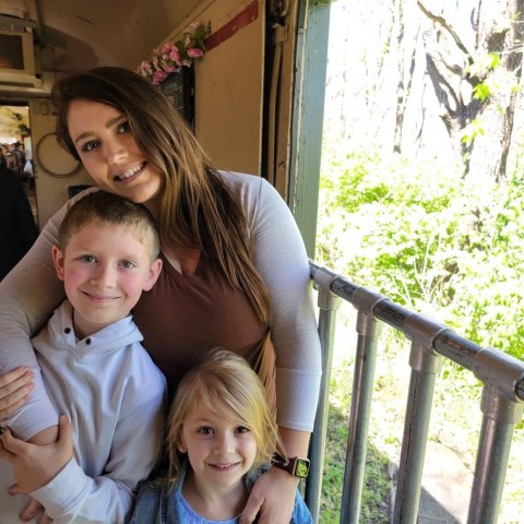 A woman hugs two smiling children on a train with a view of greenery outside.