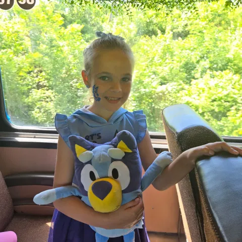 Child holding a Bluey plush toy in a train with greenery outside the window.