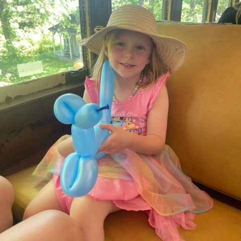 Girl in pink dress and hat holding a blue balloon animal on a train.