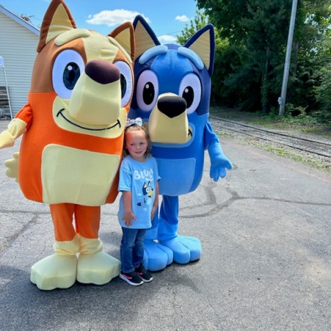 Child posing with large orange and blue dog mascots on a sunny day.