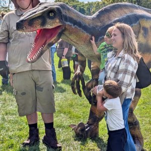 Man in safari outfit with dinosaur costume near woman and children at outdoor event.