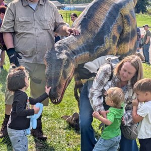 Children interact with a dinosaur puppet, guided by an adult in explorer attire in a sunny outdoor setting.