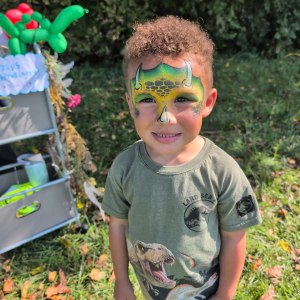 Child with dinosaur face paint and shirt standing outside on grass.